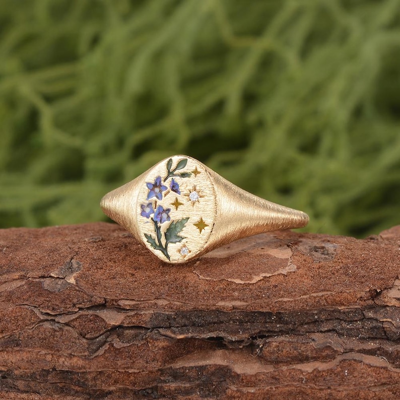 K&ouml;nnte beinhalten: Ein goldener Siegelring mit geb&uuml;rsteter Textur und ovaler Ringplatte. Die Platte zeigt ein Emaille-Design aus blauen Blumen mit gr&uuml;nen Bl&auml;ttern, akzentuiert durch kleine goldene Sterne und zwei winzige, in die Metalloberfl&auml;che eingelassene klare Steine.