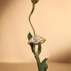 K&ouml;nnte beinhalten: Ein goldener Siegelring mit geb&uuml;rsteter Textur und ovaler Ringplatte. Die Platte ist mit einem kleinen, farbenfrohen Blumenmotiv aus violetten Bl&uuml;ten und gr&uuml;nen Bl&auml;ttern verziert, akzentuiert durch winzige, klare Steine und kleine Stern-Gravuren.