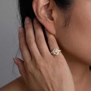 K&ouml;nnte beinhalten: Ein goldener Siegelring mit ovaler Oberfl&auml;che und eingraviertem Blumenmuster. Das florale Motiv enth&auml;lt kleine blaue und gr&uuml;ne Emaille-Akzente sowie winzige sternf&ouml;rmige Gravuren. Der Ring verf&uuml;gt &uuml;ber eine polierte Goldschiene und ein mattes Finish.