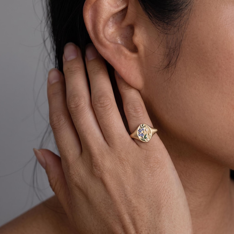 K&ouml;nnte beinhalten: Ein goldener Siegelring mit ovaler Oberfl&auml;che und eingraviertem Blumenmuster. Das florale Motiv enth&auml;lt kleine blaue und gr&uuml;ne Emaille-Akzente sowie winzige sternf&ouml;rmige Gravuren. Der Ring verf&uuml;gt &uuml;ber eine polierte Goldschiene und ein mattes Finish.
