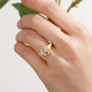 K&ouml;nnte beinhalten: Ein goldener Siegelring mit einer flachen, ovalen Oberfl&auml;che, die ein graviertes und emailliertes Design aufweist. Das Motiv zeigt kleine blaue Blumen mit gr&uuml;nen Bl&auml;ttern, akzentuiert durch winzige goldene Sterne und einen klaren Stein auf Gold.
