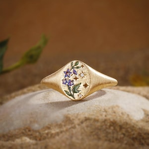 K&ouml;nnte beinhalten: Ein goldener Siegelring mit geb&uuml;rsteter Oberfl&auml;che und ovalem Kopf, verziert mit einem floralen Emaille-Design aus blauen Bl&uuml;ten und gr&uuml;nen Bl&auml;ttern, erg&auml;nzt durch kleine sternf&ouml;rmige Aussparungen und zwei winzige, eingelassene klare Steine.