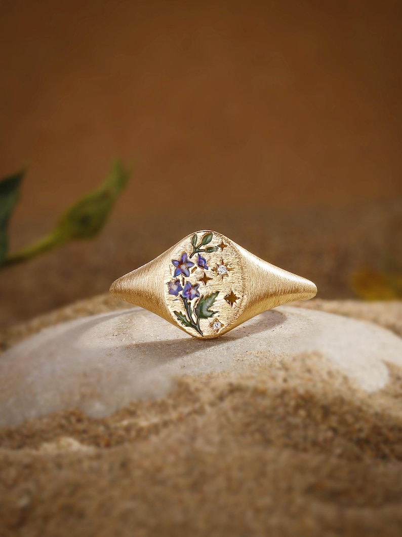 K&ouml;nnte beinhalten: Ein goldener Siegelring mit geb&uuml;rsteter Oberfl&auml;che und ovalem Kopf, verziert mit einem floralen Emaille-Design aus blauen Bl&uuml;ten und gr&uuml;nen Bl&auml;ttern, erg&auml;nzt durch kleine sternf&ouml;rmige Aussparungen und zwei winzige, eingelassene klare Steine.