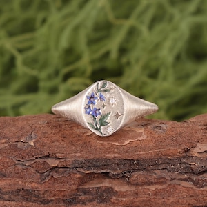 K&ouml;nnte beinhalten: Ein silberner Siegelring mit geb&uuml;rsteter Metalloberfl&auml;che und ovaler Ringplatte. Die Platte zeigt ein Emaille-Design aus blauen Blumen mit gr&uuml;nen Bl&auml;ttern und kleinen eingravierten Sternformen sowie zwei winzigen klaren Steinen.