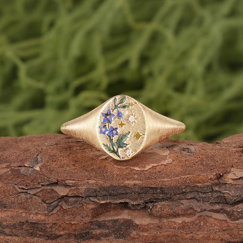 K&ouml;nnte beinhalten: Ein goldener Siegelring mit geb&uuml;rsteter Textur und einer ovalen Oberfl&auml;che, die mit blauen Emailleblumen, gr&uuml;nen Bl&auml;ttern und kleinen eingravierten Sternformen verziert ist, akzentuiert durch winzige klare Steine auf der Ringoberfl&auml;che.