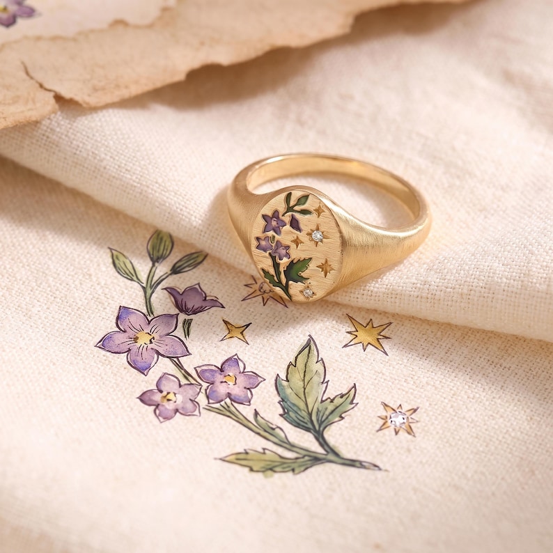 K&ouml;nnte beinhalten: Ein goldener Siegelring mit ovaler Platte, graviert mit einem Blumenmotiv aus violetten Bl&uuml;ten und gr&uuml;nen Bl&auml;ttern, verziert mit kleinen Sternformen und zwei winzigen, eingelassenen klaren Steinen auf cremefarbenem Stoff.