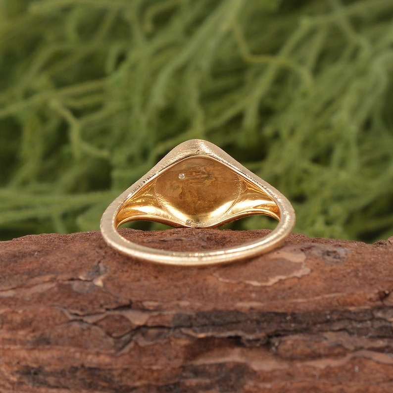 K&ouml;nnte beinhalten: Ein goldener Siegelring mit einer glatten, polierten Ringschiene und einer flachen, schildf&ouml;rmigen Oberseite. In der Mitte der Ringfl&auml;che befindet sich ein kleiner, einzelner, klarer Stein, der einen Lichtpunkt auf der goldenen Oberfl&auml;che bildet.
