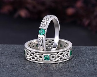 May include: Two silver rings with Celtic knot designs and emerald green gemstones. One ring features a single emerald, while the other has multiple emeralds set within the intricate knotwork. The rings are displayed on a dark gray surface.