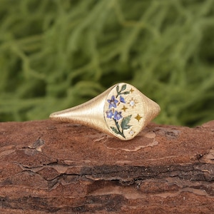 K&ouml;nnte beinhalten: Ein goldener Siegelring mit geb&uuml;rsteter Textur und ovaler Ringplatte. Die Platte zeigt ein gemaltes Motiv aus blauen Blumen mit gr&uuml;nen Bl&auml;ttern, akzentuiert durch kleine goldene Sternformen und zwei winzige klare Steine in der Oberfl&auml;che.