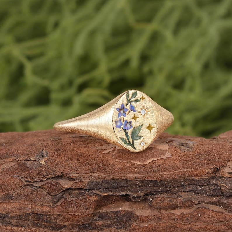 K&ouml;nnte beinhalten: Ein goldener Siegelring mit geb&uuml;rsteter Textur und ovaler Ringplatte. Die Platte zeigt ein gemaltes Motiv aus blauen Blumen mit gr&uuml;nen Bl&auml;ttern, akzentuiert durch kleine goldene Sternformen und zwei winzige klare Steine in der Oberfl&auml;che.