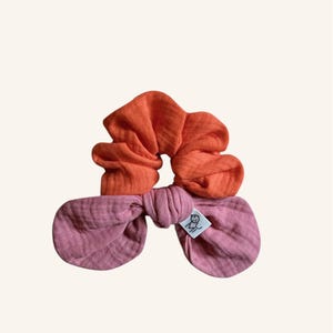 Puede incluir: Dos coleteros. El superior es naranja, el inferior rosa y con forma de lazo. Una pequeña etiqueta blanca con un logotipo está adherida al coletero rosa. Los coleteros están hechos de una tela suave.