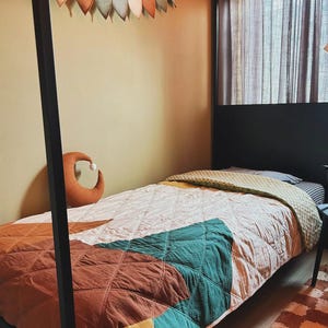 Puede incluir: Una cama con estructura de metal negro y una colcha acolchada con bloques geométricos en tonos óxido, verde azulado y amarillo mostaza. Un cojín en forma de media luna color óxido descansa sobre la cama y una guirnalda de tela cuelga arriba.