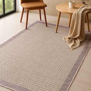Puede incluir: Una alfombra rectangular con un patrón tejido en tonos neutros, enmarcada por un borde lavanda. La alfombra está colocada sobre un suelo de madera clara, con una silla marrón y una mesa pequeña en el fondo.