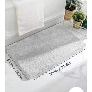 Op de afbeelding: Een rechthoekige grijze badmat, 80 cm lang en 50 cm breed, ligt op een betegelde vloer. De mat heeft een zacht, getextureerd oppervlak. Een witte handdoek hangt over de rand van een witte badkuip.