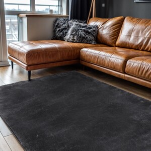 May include: A dark gray rectangular area rug on a hardwood floor. The rug is in front of a brown leather sectional sofa with two fluffy gray pillows. A wooden floor lamp with a black shade is in the background.