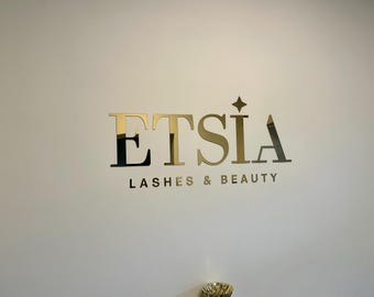 May include: Gold metallic sign on a white wall. The sign reads "ETSIA" with "LASHES & BEAUTY" below. A star is above the letter "A". The sign is likely for a beauty salon or spa.