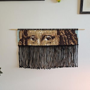 Op de afbeelding: Een macramé wandkleed met een gedetailleerd portret van een gezicht. Het kunstwerk gebruikt tinten bruin, beige en zwart om de afbeelding te creëren, met een lichtblauwe rand. Zwarte franjes hangen van de onderkant en het stuk is gemonteerd op een houten deuvel.