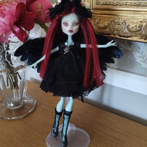 Peut inclure: Une poupée personnalisée avec une peau pâle, des cheveux roux et une robe noire. La poupée a des ailes noires en plumes et porte des bottes noires. Un coiffe floral noir orne la tête de la poupée. La poupée est sur un support en plastique transparent.
