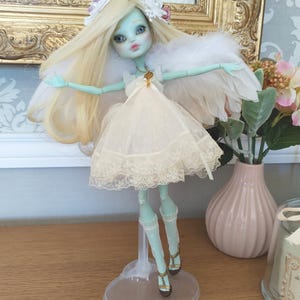 Peut inclure: Une poupée personnalisée à la peau vert pâle, aux longs cheveux blonds et à une robe blanche avec une bordure en dentelle. La poupée a des ailes en plumes et un serre-tête floral. Elle est sur un support en plastique transparent, mettant en valeur un design unique et artisanal.