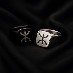 May include: Two square-faced rings with a geometric design. One ring is black with a silver design, and the other is silver with a black design. The rings are displayed on a black fabric background.