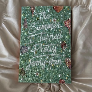 Könnte beinhalten: Ein Buch mit dem Titel "The Summer I Turned Pretty" von Jenny Han. Das Buchcover ist grün mit dem Titel in weißer Schrift und dekorativen Elementen. Das Buch liegt auf einem cremefarbenen, strukturierten Stoff.