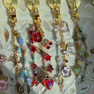 Pode incluir: Seis chaveiros decorativos com ferragens douradas e fechos em forma de coração. Cada chaveiro apresenta um design único com contas, berloques e pingentes coloridos, incluindo flores, borboletas e corações. Os chaveiros são exibidos em uma superfície decorativa.