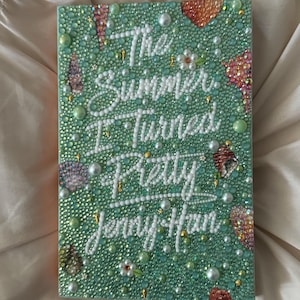 Puede incluir: Una portada del libro "El verano en que me enamoré" de Jenny Han. La portada es de color verde mar y está cubierta de pequeños adornos redondos y brillantes. El título y el nombre del autor están escritos en blanco.