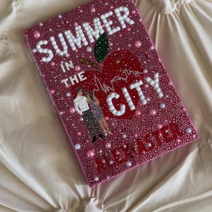 以下が含まれることがあります： 白い真珠のような文字で「Summer in the City」と書かれたピンク色の装飾が施された本の表紙。都市のスカイラインを備えた赤いリンゴのデザインが特徴です。表紙にはカップルのイラストも含まれています。