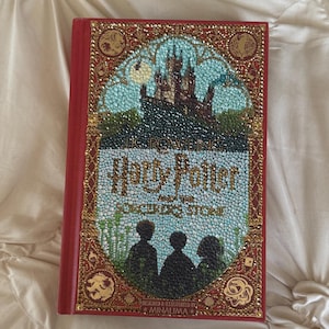 May include: A red hardcover book titled "Harry Potter and the Sorcerer's Stone." The cover features a detailed illustration of a castle, silhouettes, and the book title in a decorative font. The cover is embellished with gold accents.
