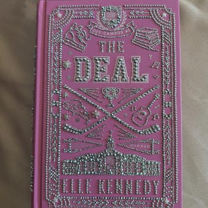 K&ouml;nnte beinhalten: Rosa Hardcover-Buch mit dem Titel "The Deal" von Elle Kennedy. Das Cover ist mit silberfarbenen Strasssteinen verziert, die dekorative R&auml;nder, Text und Illustrationen von Sportger&auml;ten und einem Geb&auml;ude bilden.