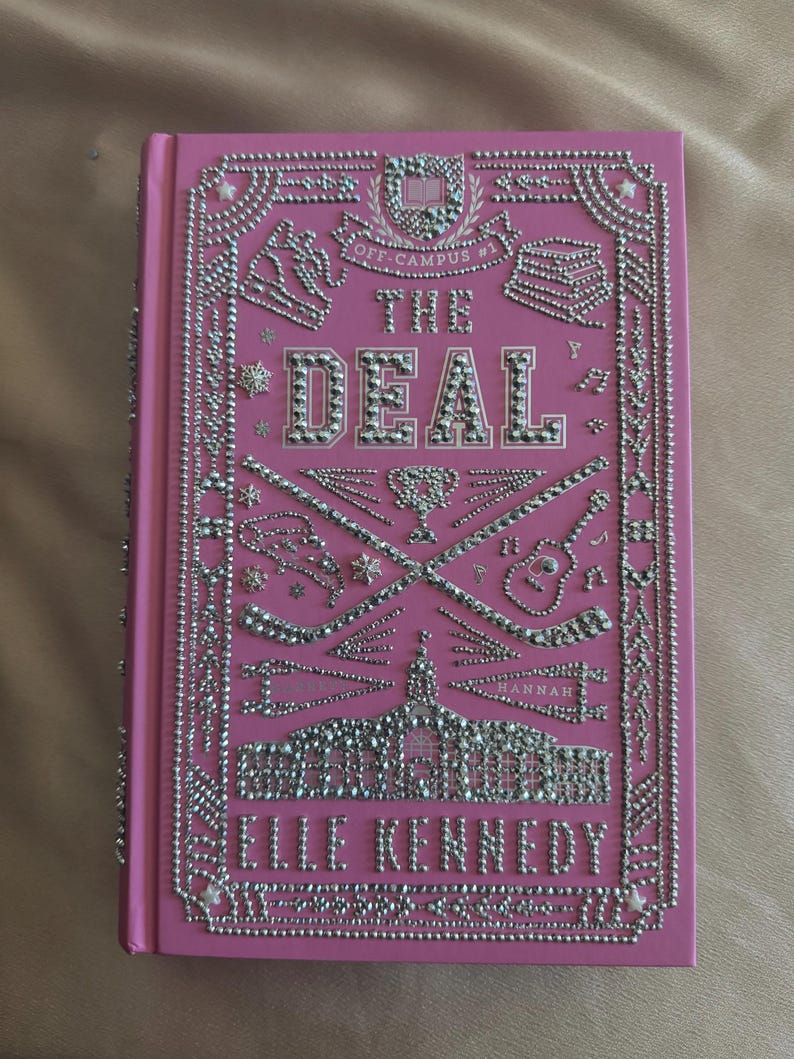 K&ouml;nnte beinhalten: Rosa Hardcover-Buch mit dem Titel "The Deal" von Elle Kennedy. Das Cover ist mit silberfarbenen Strasssteinen verziert, die dekorative R&auml;nder, Text und Illustrationen von Sportger&auml;ten und einem Geb&auml;ude bilden.