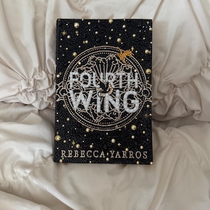 May include: Hardcover book titled "Fourth Wing" by Rebecca Yarros. The book has a black cover adorned with gold and silver embellishments, including a circular design and small stars. The book is resting on a cream-colored, textured surface.