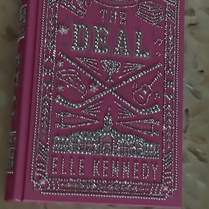 K&ouml;nnte beinhalten: Ein pinkfarbenes Hardcover-Buch mit dem Titel "THE DEAL" von Elle Kennedy. Das Cover zeigt silberne Verzierungen und dekorative Elemente. Der Titel und der Name des Autors sind in silbernen Buchstaben gehalten.