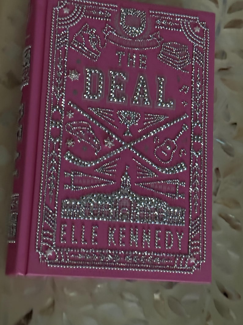 K&ouml;nnte beinhalten: Ein pinkfarbenes Hardcover-Buch mit dem Titel "THE DEAL" von Elle Kennedy. Das Cover zeigt silberne Verzierungen und dekorative Elemente. Der Titel und der Name des Autors sind in silbernen Buchstaben gehalten.
