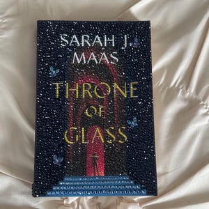 May include: A hardcover book titled "Throne of Glass" by Sarah J. Maas. The cover is dark blue with sparkling embellishments and features the title in gold lettering. A silhouette of a person stands before a red archway.