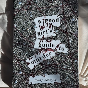 以下が含まれることがあります： ホリー・ジャクソン著「A Good Girl's Guide to Murder」というタイトルの本。表紙は銀色のラインストーンと赤い糸で装飾されており、タイトルと著者の名前はかすれたフォントで表示されています。