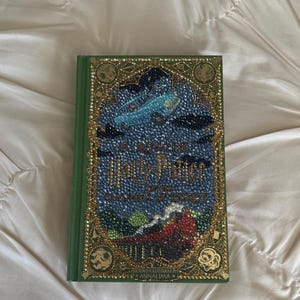 Può includere: Un libro con copertina rigida intitolato Harry Potter and the Chamber of Secrets, progettato e illustrato da Minalima. La copertina verde presenta un bordo dorato decorativo e una scena centrale realizzata con strass colorati, raffigurante un'auto volante.