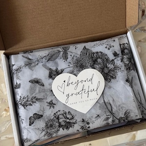 K&ouml;nnte beinhalten: Ein offener Pappkarton mit wei&szlig;em Seidenpapier mit einem schwarz-wei&szlig;en Blumen- und Schmetterlingsmuster. Ein wei&szlig;er herzf&ouml;rmiger Aufkleber mit den Worten "beyond grateful" liegt oben auf dem Seidenpapier.