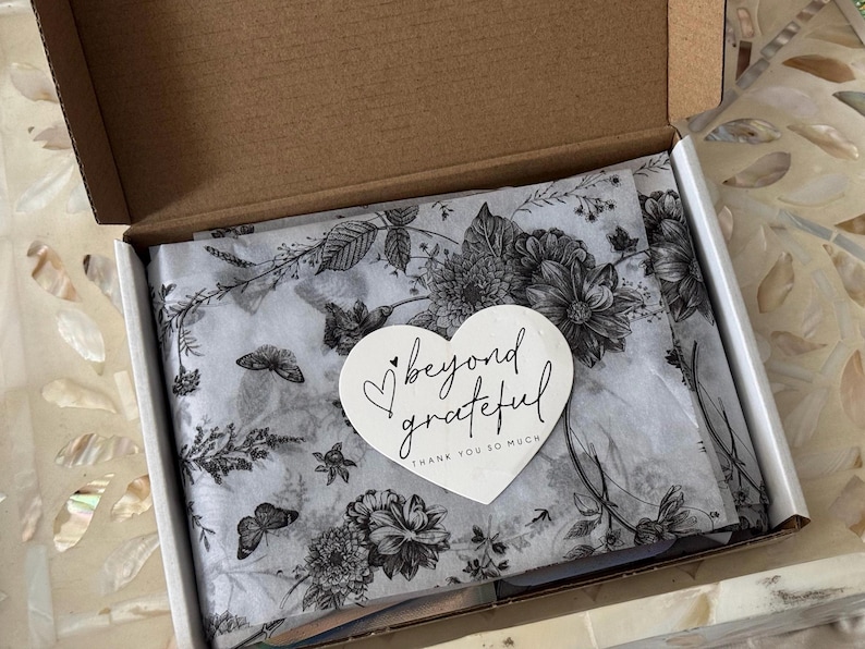 K&ouml;nnte beinhalten: Ein offener Pappkarton mit wei&szlig;em Seidenpapier mit einem schwarz-wei&szlig;en Blumen- und Schmetterlingsmuster. Ein wei&szlig;er herzf&ouml;rmiger Aufkleber mit den Worten "beyond grateful" liegt oben auf dem Seidenpapier.