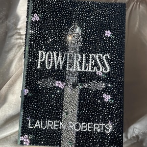 以下が含まれることがあります： ローレン・ロバーツ著「POWERLESS」というタイトルの黒いハードカバーの本。キラキラ光る装飾が施されています。銀色の十字のデザインが表紙の中央にあり、小さな紫色の花が散りばめられています。