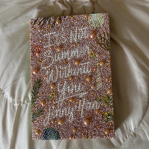 May include: A rectangular, pink, bedazzled art piece with the text "It's Not Summer Without You Jenny Han" in white script. The piece is covered in small, iridescent rhinestones and pearl-like beads, with accents of green and gold.