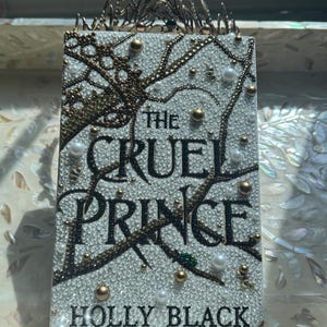 K&ouml;nnte beinhalten: Buchcover von "The Cruel Prince" von Holly Black, verziert mit Kristallen, Perlen und goldenen Akzenten. Titel und Autorenname sind in Schwarz gehalten. Eine dekorative Krone befindet sich oben auf dem Buch.
