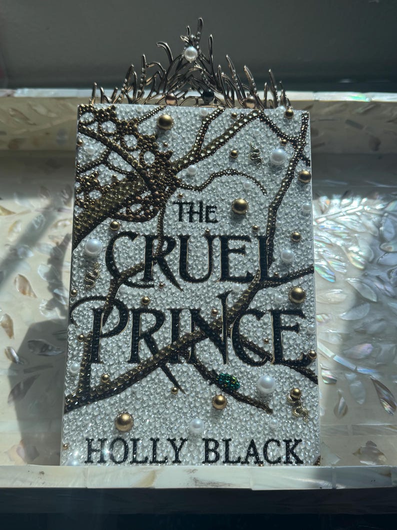 K&ouml;nnte beinhalten: Buchcover von "The Cruel Prince" von Holly Black, verziert mit Kristallen, Perlen und goldenen Akzenten. Titel und Autorenname sind in Schwarz gehalten. Eine dekorative Krone befindet sich oben auf dem Buch.