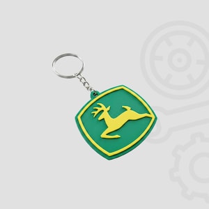 Puede incluir: Un llavero verde y amarillo con un emblema de ciervo. El llavero tiene un fondo verde con un ciervo amarillo y un borde amarillo. El llavero está unido a un anillo y una cadena de metal plateado.