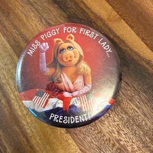 May include: A round button featuring Miss Piggy from The Muppets, with the text "MISS PIGGY FOR FIRST LADY... PRESIDENT!" The image shows Miss Piggy in a pink dress and purple gloves, with American flags in the background.