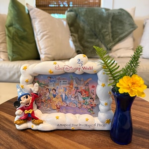 May include: A white cloud-shaped Walt Disney World 25th anniversary picture frame featuring Mickey Mouse in a sorcerer outfit, yellow stars, and a colorful illustration of Disney characters with the text Remember the Magic and A Magical Time in a Magical Place.