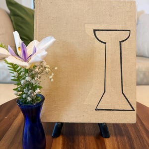 May include: A beige canvas art piece with a black geometric design, displayed on a small black easel. A small blue vase holds a white and purple flower arrangement, set on a wooden table.