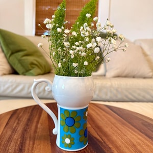 May include: A white ceramic mug with a handle, decorated with a blue and green floral pattern, holding a bouquet of white flowers and green foliage. The mug sits on a wooden table.