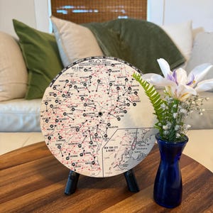 May include: A round Dist-O-Map on a black stand, displaying a vintage map with red and black details. A small blue vase with white and purple flowers sits nearby on a wooden table. The map shows cities like New York and Boston.