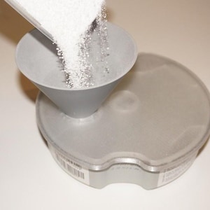 May include: A grey plastic funnel pouring white granules into a round, grey container. The container has a slightly textured surface and a label with a barcode. The image is taken from a high angle.